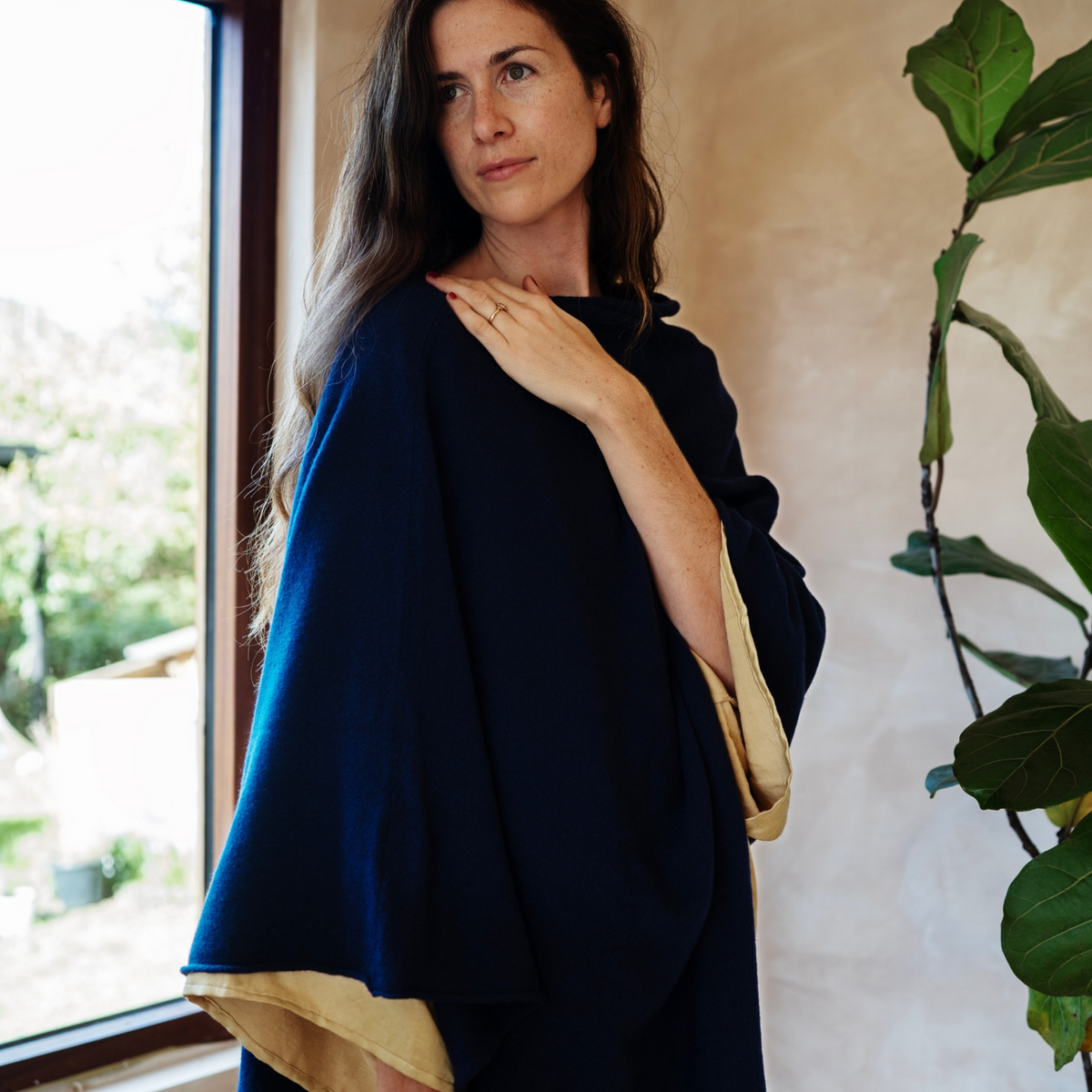 A woman wearing a dark navy blue wrap or poncho with a contrasting beige lining, standing in a softly lit room next to a window and a potted plant.