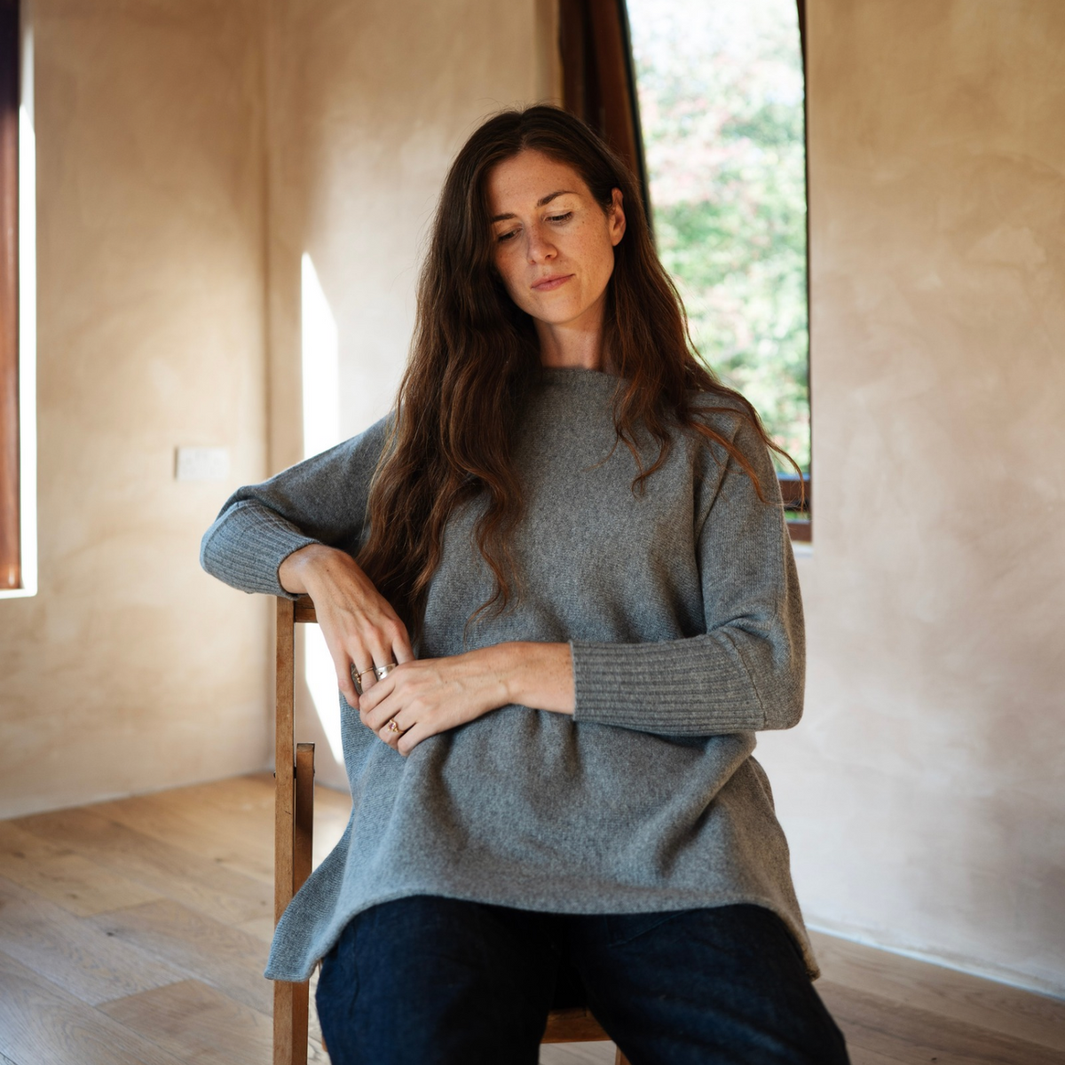 A woman with long brown hair sits on a wooden chair in a rustic room, wearing a loose-fit gray knit sweater and dark jeans.