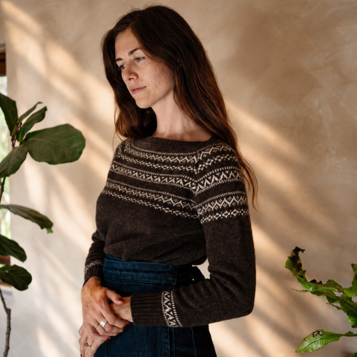 A woman wearing a dark brown knit sweater with a cream geometric pattern, paired with a blue denim skirt, standing in a sunlit room with plants and a textured beige wall.