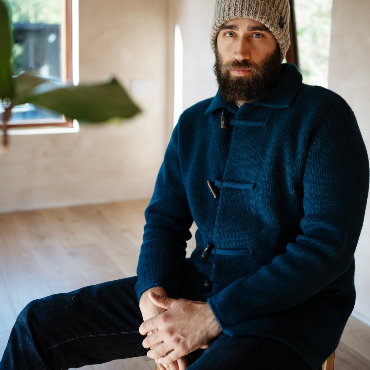 A man with a beard wearing a dark navy blue wool duffle coat and a beige knit beanie, sitting in a softly lit indoor room with light wood floors and a window in the background.