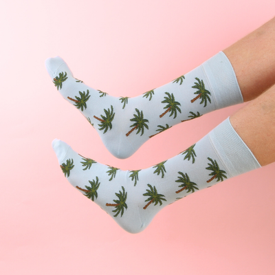 Pale Blue socks with palm tree pattern worn by a person on a pink background