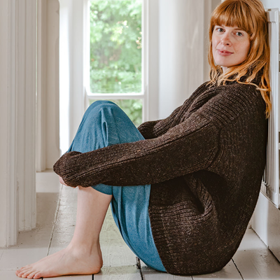 Woman sitting on the floor wearing a large brown knitted sweater.