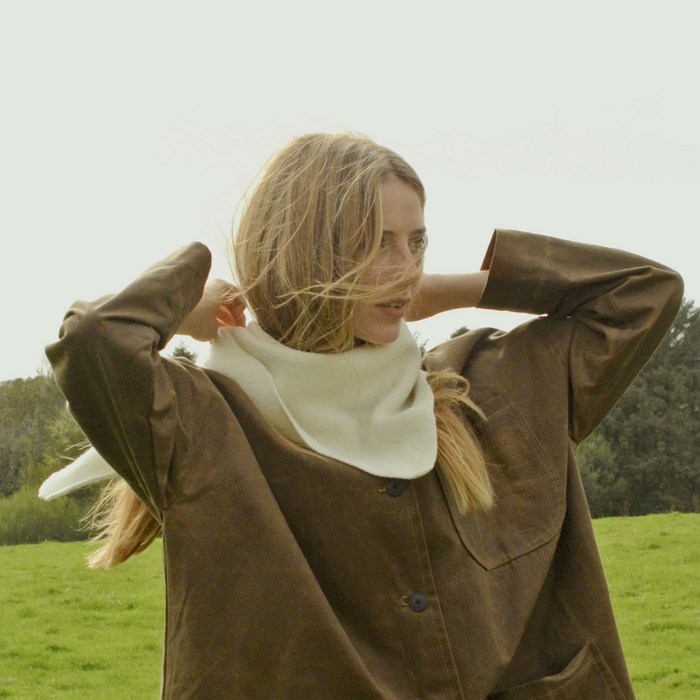 Woman in a brown coat standing in a grassy field with trees in the background wearing a white triangular scarf