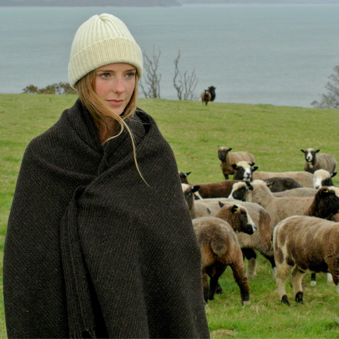 Woman wrapped in a blanket and hat standing in a field with sheep