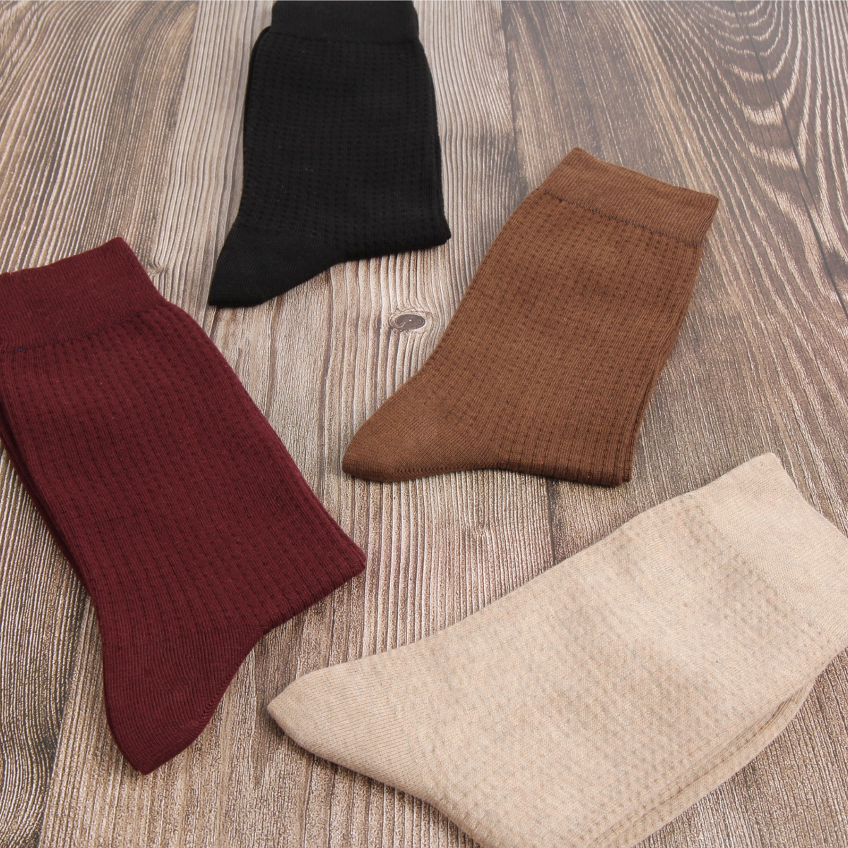 Four pairs of ribbed waffle-knit ankle socks in black, brown, burgundy, and beige, arranged on a light brown wood grain background.