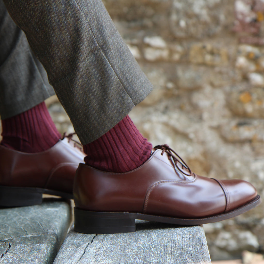 A close-up of a person's legs wearing brown leather cap-toe oxford shoes, burgundy ribbed socks, and gray herringbone trousers, standing on a weathered wooden surface with a blurred stone wall background.