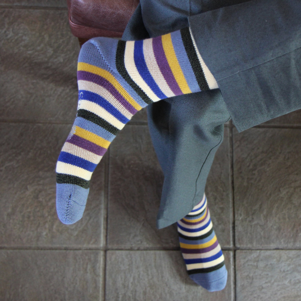 A person wearing blue striped socks with purple, yellow, white, and black bands, paired with blue trousers, sitting on a tiled floor.