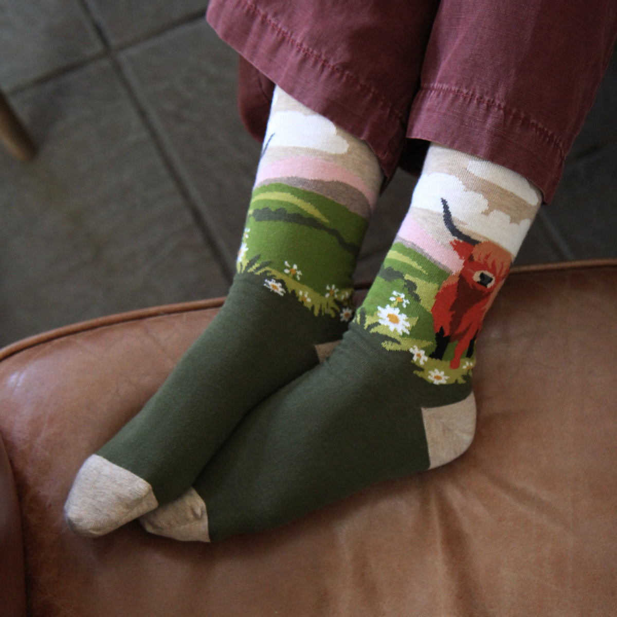 A person sitting on a brown leather chair wearing dark olive green socks with a scenic landscape print on the upper leg area, featuring green hills, pink clouds, white daisies, and a red cow. The person is also wearing maroon pants.