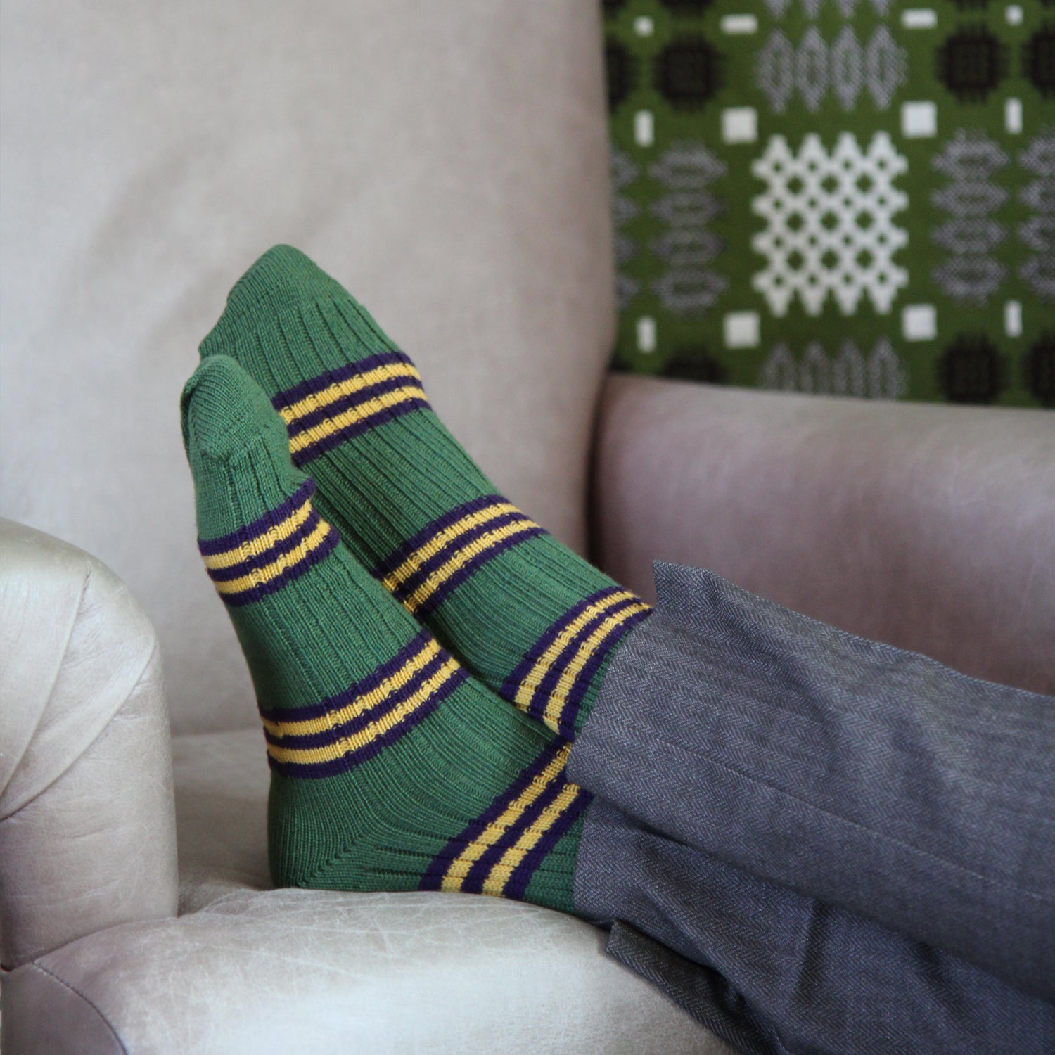 A pair of green knit socks with yellow and purple stripes, worn on feet resting on a beige armchair.