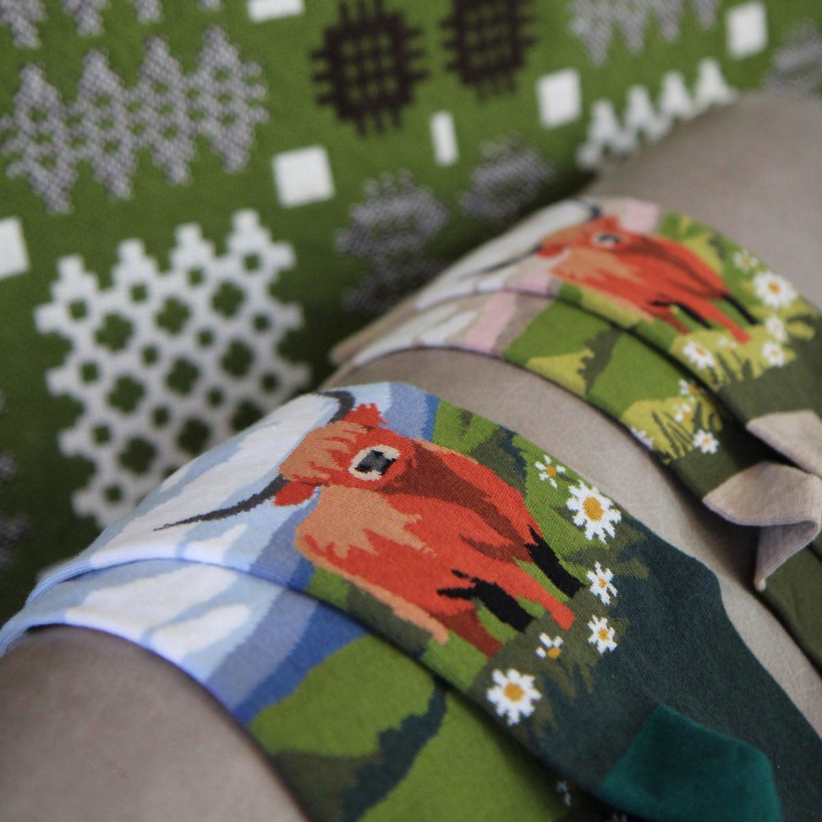 A close-up of a person's leg wearing a pair of colorful knit socks with a red goat, white daisies, and blue landscape print, next to a green patterned knit blanket.