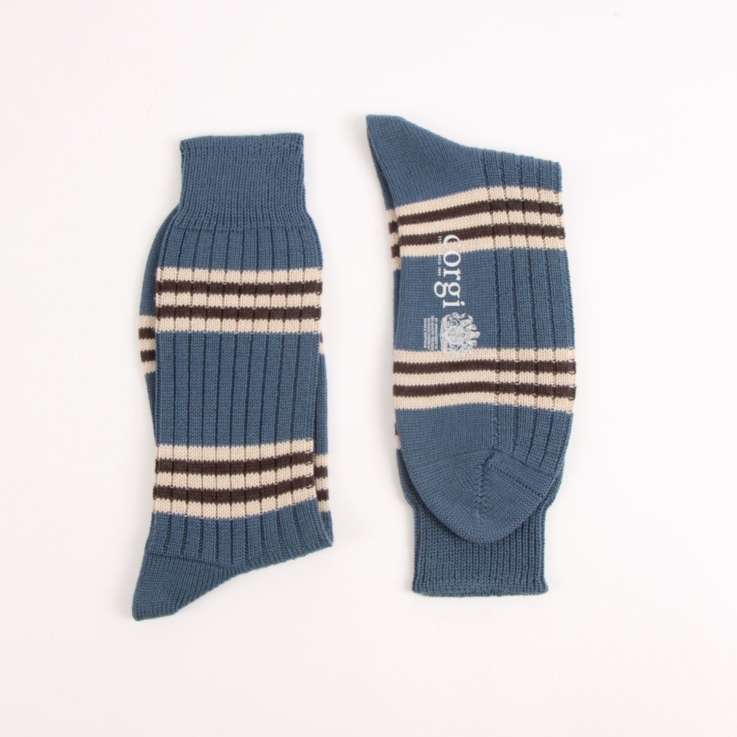 A pair of ribbed knit crew socks in a muted slate blue color, featuring horizontal stripes in cream and dark brown, with a white printed logo on the side.