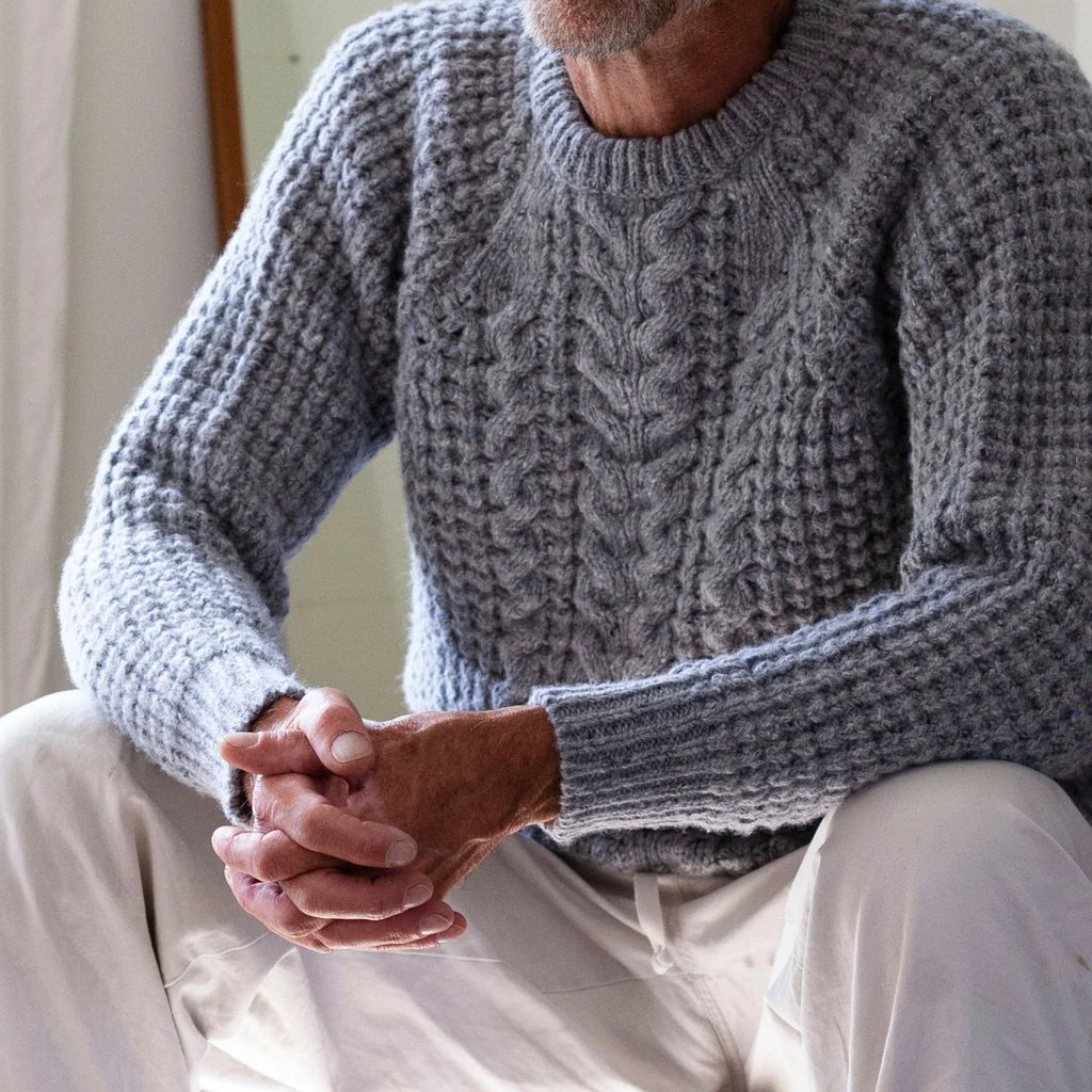 A close-up of a man wearing a chunky gray cable-knit sweater and light-colored pants, sitting indoors with his hands clasped.