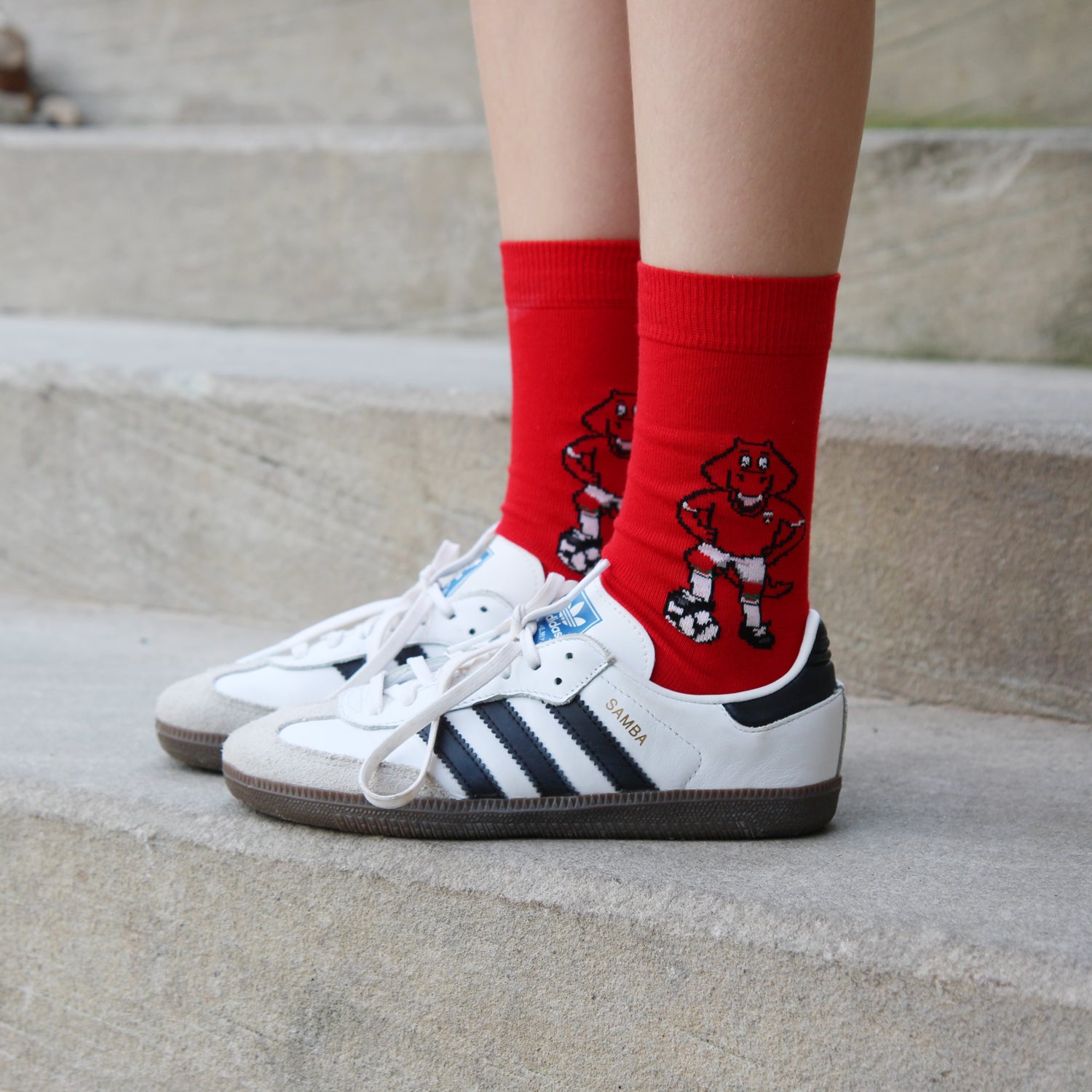Red crew socks with a cartoon character graphic, worn with white Adidas Samba sneakers on concrete steps.
